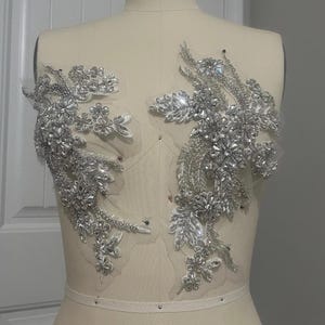 May include: A pair of silver beaded appliques with a floral design. The appliques are on a white mesh background.