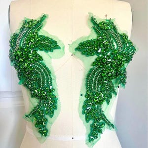 May include: Two green beaded appliques with a leaf design. The appliques are made of green beads and sequins, and are attached to a sheer, light green fabric backing. The appliques are displayed on a mannequin, likely for dressmaking.