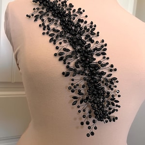May include: A black beaded and leaf-shaped hair accessory. The accessory is made of small, round beads and leaf-shaped accents, arranged in a flowing, asymmetrical design. The piece is attached to a wire frame, creating a delicate and elegant look.