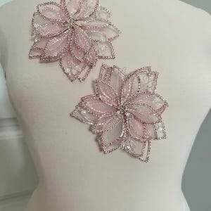 Pode incluir: Duas aplicações florais rosa com strass e contas transparentes e rosa. As aplicações são feitas de pétalas transparentes em camadas e são presas a um manequim. As aplicações são provavelmente para embelezar roupas ou acessórios.