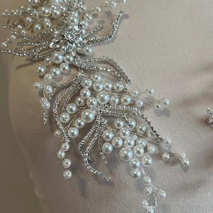 May include: A silver and pearl bridal applique. The design features clusters of white pearls and sparkling rhinestones arranged in a floral pattern. The applique is designed to be sewn onto fabric.