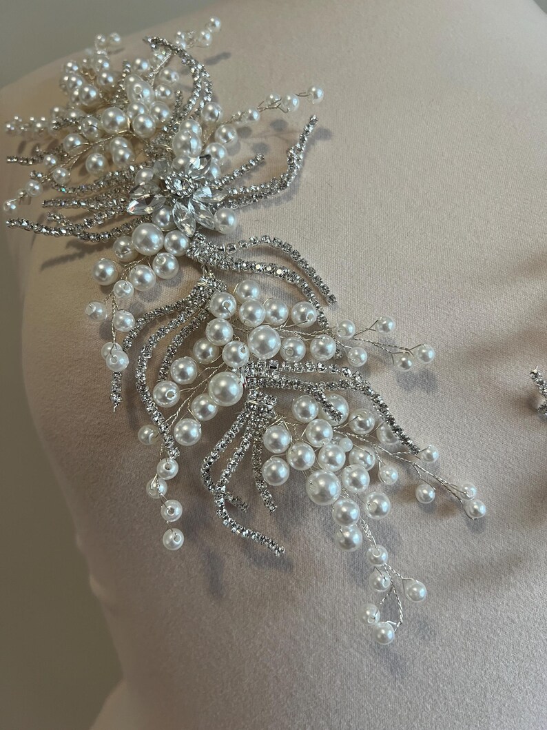 May include: A silver and pearl bridal applique. The design features clusters of white pearls and sparkling rhinestones arranged in a floral pattern. The applique is designed to be sewn onto fabric.
