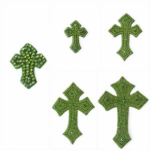 May include: A collection of five green rhinestone crosses of varying sizes. Each cross is covered in small, sparkling green rhinestones, with a detailed design. The crosses are displayed against a white background.