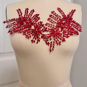 K&ouml;nnte beinhalten: Rote, perlenbesetzte Applikation mit floralem Design und blattartigen Akzenten. Die Applikation wird auf einer Schneiderpuppe pr&auml;sentiert, um ihre Form und m&ouml;gliche Verwendung zur Verzierung von Kleidungsst&uuml;cken oder Accessoires zu zeigen.