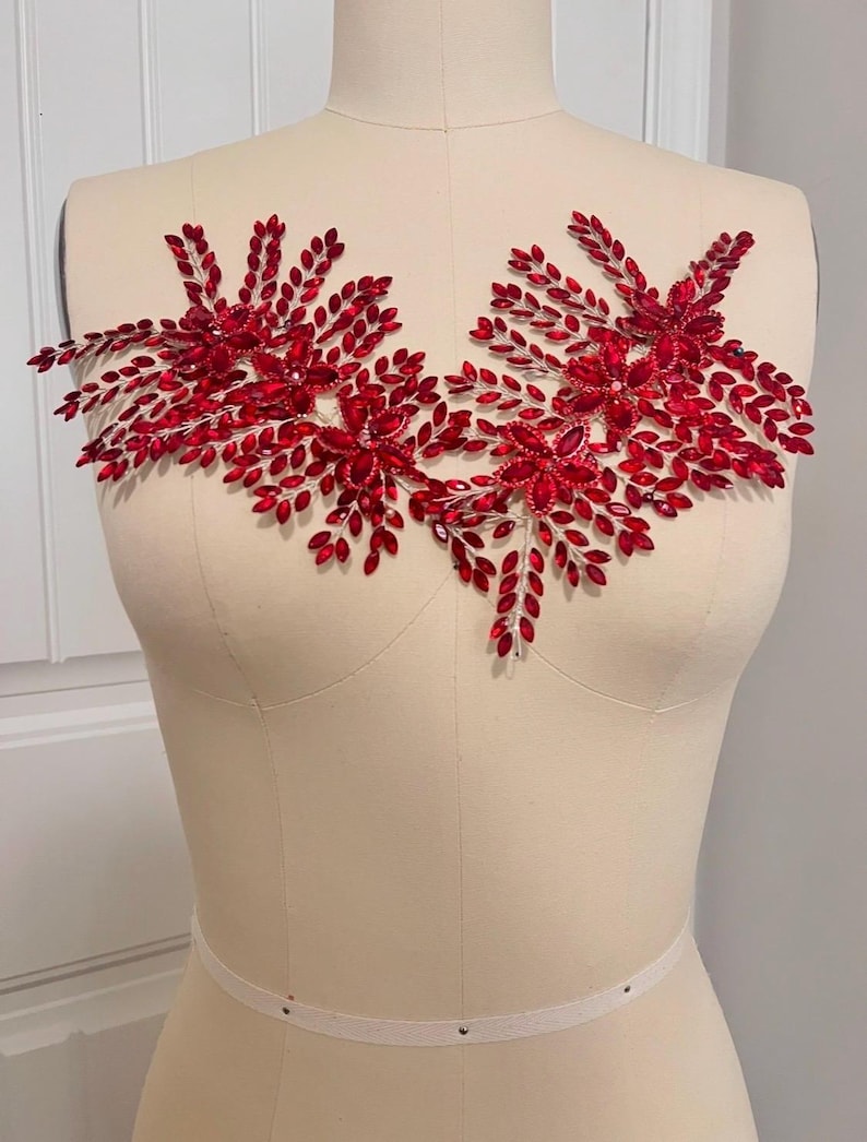 K&ouml;nnte beinhalten: Rote, perlenbesetzte Applikation mit floralem Design und blattartigen Akzenten. Die Applikation wird auf einer Schneiderpuppe pr&auml;sentiert, um ihre Form und m&ouml;gliche Verwendung zur Verzierung von Kleidungsst&uuml;cken oder Accessoires zu zeigen.