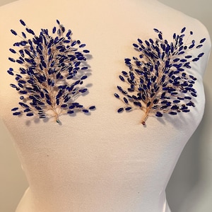 May include: Two decorative floral hair accessories. Each features a gold-toned wire base with clusters of dark blue beads, resembling stylized branches. The accessories are displayed on a white mannequin, showcasing their design and form.