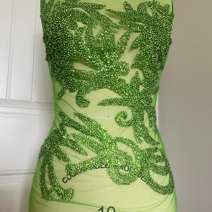 May include: Green mesh fabric with a large green rhinestone design. The design is a stylized floral pattern. The fabric is on a dress form.