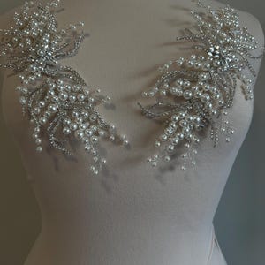 May include: Two silver-toned, floral-shaped embellishments. Each features a central cluster of clear rhinestones surrounded by delicate silver wire, faux pearls, and smaller rhinestones. These decorative pieces are likely for bridal or formal wear.