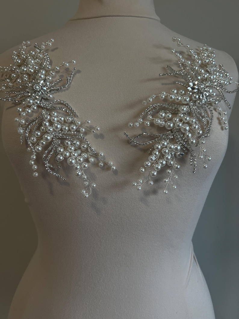 May include: Two silver-toned, floral-shaped embellishments. Each features a central cluster of clear rhinestones surrounded by delicate silver wire, faux pearls, and smaller rhinestones. These decorative pieces are likely for bridal or formal wear.