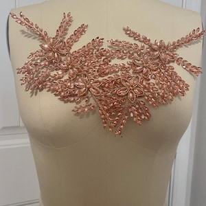 K&ouml;nnte beinhalten: Ros&eacute;goldfarbene Perlenapplikation mit floralem Design. Die Applikation weist Cluster aus funkelnden Kristallen und blattf&ouml;rmigen Perlen auf, wodurch eine zarte und verzierte Verzierung entsteht. Das Design ist symmetrisch.