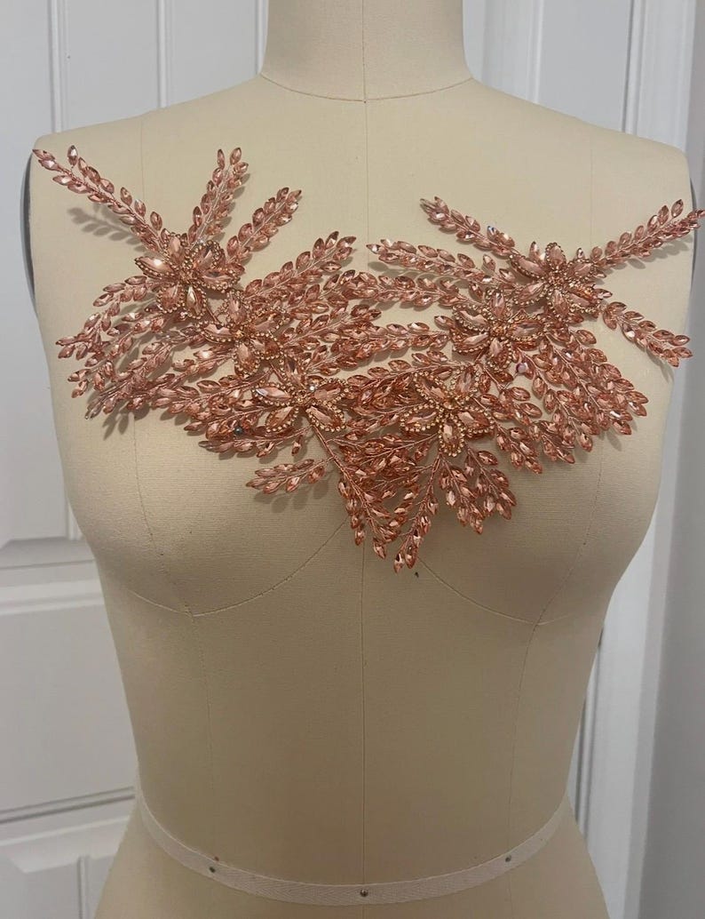 K&ouml;nnte beinhalten: Ros&eacute;goldfarbene Perlenapplikation mit floralem Design. Die Applikation weist Cluster aus funkelnden Kristallen und blattf&ouml;rmigen Perlen auf, wodurch eine zarte und verzierte Verzierung entsteht. Das Design ist symmetrisch.