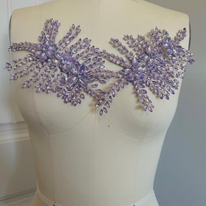 K&ouml;nnte beinhalten: Zwei symmetrische, floral inspirierte Verzierungen in hellem Lila. Jedes Design zeigt Cluster aus kleinen, blattf&ouml;rmigen Kristallen und gr&ouml;&szlig;eren, facettierten Kristallen. Die Verzierungen werden auf einer Schaufensterpuppe pr&auml;sentiert.
