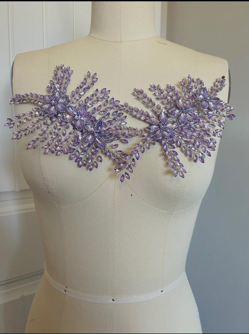 K&ouml;nnte beinhalten: Zwei symmetrische, floral inspirierte Verzierungen in hellem Lila. Jedes Design zeigt Cluster aus kleinen, blattf&ouml;rmigen Kristallen und gr&ouml;&szlig;eren, facettierten Kristallen. Die Verzierungen werden auf einer Schaufensterpuppe pr&auml;sentiert.