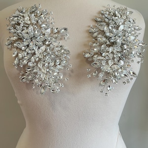 May include: Two silver crystal embellishments, each featuring a cluster of various-sized clear crystals and small pearls. The crystals are arranged in a floral pattern, with delicate wire stems. The embellishments are displayed on a mannequin.