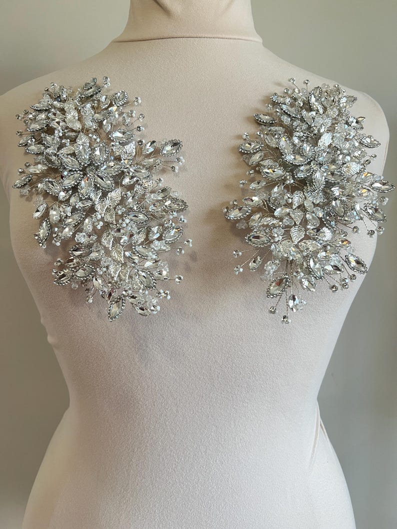 May include: Two silver crystal embellishments, each featuring a cluster of various-sized clear crystals and small pearls. The crystals are arranged in a floral pattern, with delicate wire stems. The embellishments are displayed on a mannequin.