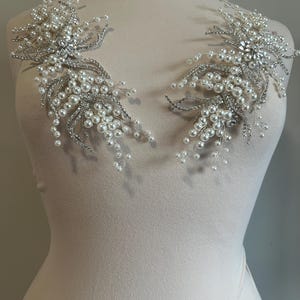 May include: Two silver and pearl shoulder embellishments. Each piece features a central cluster of clear rhinestones surrounded by a spray of faux pearls and silver wire. The design is symmetrical, with each piece mirroring the other.