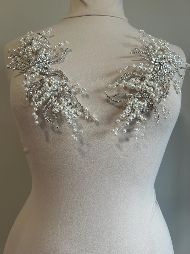 May include: Two silver and pearl shoulder embellishments. Each piece features a central cluster of clear rhinestones surrounded by a spray of faux pearls and silver wire. The design is symmetrical, with each piece mirroring the other.