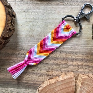 Può includere: Un portachiavi a chevron rosa, arancione, bianco e giallo con un anello portachiavi argentato. Il portachiavi ha un fiocco all'estremità.