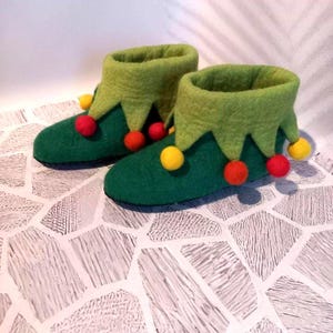 May include: A pair of green felt elf boots with a light green collar and red and yellow pom-poms. The boots have a pointed collar and are designed to look like elf shoes. The boots are on a white and gray patterned surface.