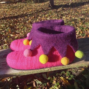 May include: A pair of pink felt slippers with a purple jester-style collar. The slippers are adorned with yellow, orange, and pink pom-poms. The slippers are sitting on a wooden surface.