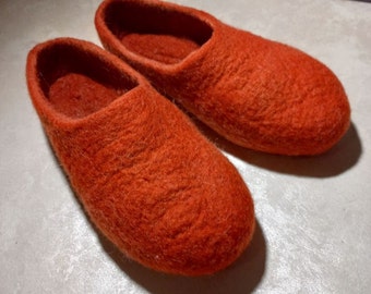 May include: A pair of orange felted wool slippers. The slippers have a rounded toe and a low-cut design, showcasing a textured, fuzzy surface. The slippers are designed for indoor use and are placed on a light-colored surface.