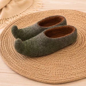 May include: A pair of green felt slippers with upturned toes and brown interior lining. The slippers are displayed on a round, woven mat. The felt has a textured appearance, and the slippers appear handmade.