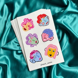 May include: A stack of stickers featuring colorful, stylized faces with various hairstyles and expressions. The stickers are on a white background, and the text "@meet_santa" is visible. The background is a teal satin fabric.