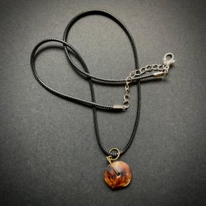 Può includere: Una collana artigianale con un ciondolo rotondo naturale con toni marroni e beige, avvolto in filo dorato. Il ciondolo pende da una collana di corda nera con chiusura argentata e catena regolabile.