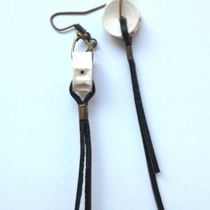 Può includere: Un paio di orecchini con ciondoli in ceramica bianca e nappe in pelle nera. I ciondoli sono di forme diverse, uno è quadrato e l'altro è un cerchio.
