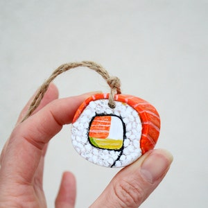 May include: A hand-painted sushi roll ornament with a twine loop. The ornament has a salmon-coloured top, white rice, and a colourful filling of yellow, orange, and green. A unique, food-themed decoration.