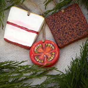 May include: Three hanging ornaments featuring food-themed designs: a square white piece with a red stripe, a square brown textured piece, and a round red slice showing the interior seeds and structure of a tomato, each with a jute twine loop.