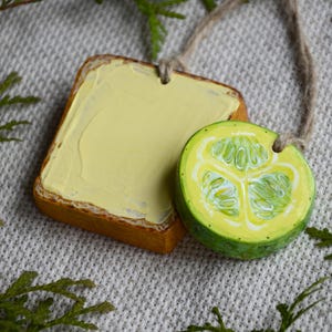 May include: Two hanging ornaments shaped like food items. One is a square piece of toast topped with a thick layer of pale yellow butter. The other is a round slice of a green cucumber with visible seeds, both attached to a brown twine string.