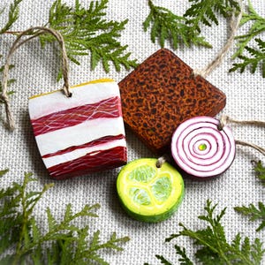 May include: Four food-themed ornaments featuring a rectangular piece of bacon with red and white stripes, a square brown textured piece, a round purple and white onion slice, and a round green lime slice, each with a jute twine hanging loop.