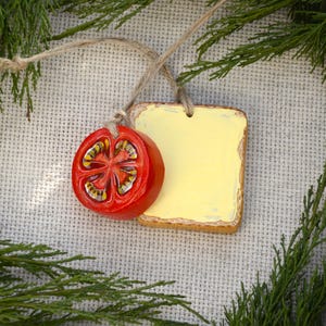 May include: A decorative ornament featuring a square piece of toast with a thick layer of yellow butter and a bright red tomato slice. Both pieces are crafted from a smooth material and are suspended together by a thin, natural-colored twine string.