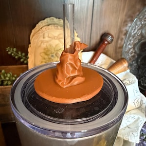 May include: A brown tumbler lid with a Sorting Hat design and a clear straw. The lid is set on a silver tumbler. The Sorting Hat is a detailed, three-dimensional design. The tumbler is likely for cold beverages.