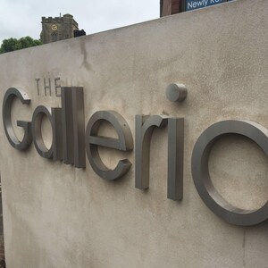 May include: A beige concrete wall featuring the three-dimensional metallic silver text The Galleria. The letters are mounted to the surface, with a small circular dot above the letter i. The sign is positioned outdoors in an urban setting.