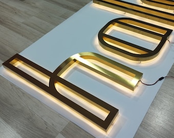 May include: Gold-colored metal letters spelling "hello" with illuminated edges. The letters are mounted on a white surface and connected to electrical wires.