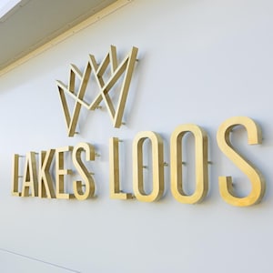 K&ouml;nnte beinhalten: Goldfarbenes Metallschild mit der Aufschrift "LAKES LOOS" und einem kronenartigen Emblem dar&uuml;ber. Das Schild ist an einer wei&szlig;en Wand befestigt. Die Buchstaben sind dick und dreidimensional, was ein klares Statement ergibt.