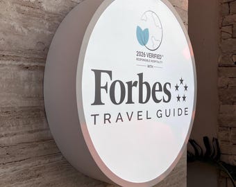 Op de afbeelding: Een rond, verlicht wandbord met het Forbes Travel Guide logo in donkergrijze tekst. Boven de tekst staat een afbeelding van een wereldbol met twee lichtblauwe bladeren en de tekst 2026 VERIFIED RESPONSIBLE HOSPITALITY.