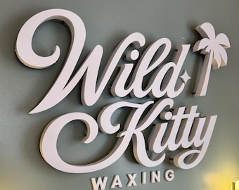 May include: A three-dimensional wall sign featuring the white text Wild Kitty in a cursive script font, with a small white palm tree icon and a four-pointed star. Below the cursive text, the word WAXING is displayed in a simple sans-serif font.