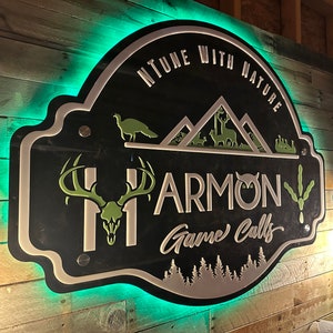 May include: A black, wall-mounted sign with green LED backlighting featuring the text "NTUNE WITH NATURE" and "HARMON Game Calls". The design includes a deer skull, an owl, a turkey, mountain silhouettes, and pine trees in white and green.