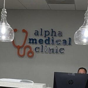 May include: A reception desk with a wavy dark brown front panel and a light-colored countertop. On the gray wall behind the desk, a sign reads alpha medical clinic in dark letters with an orange stethoscope graphic. Two glass pendant lights hang above.