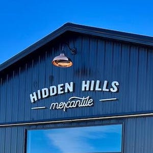 May include: Exterior view of a dark blue building with the text "HIDDEN HILLS - mercantile" in white. The building has a large glass door and window, with a clear blue sky in the background.