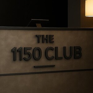 May include: A rectangular reception desk with a matte dark gray finish and a black base. The front features raised black lettering that reads THE 1150 CLUB with a horizontal line underneath. A small lamp with a warm white shade sits to the right.