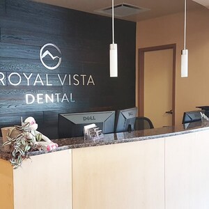 May include: A dental office reception desk featuring a light wood front, a dark granite countertop, and a black wood-paneled wall behind it. The wall displays the silver text ROYAL VISTA DENTAL with a circular mountain logo above the lettering.