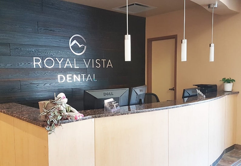 May include: A dental office reception desk featuring a light wood front, a dark granite countertop, and a black wood-paneled wall behind it. The wall displays the silver text ROYAL VISTA DENTAL with a circular mountain logo above the lettering.
