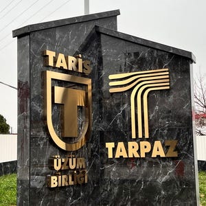 Puede incluir: Un gran monumento angular hecho de m&aacute;rmol oscuro pulido con vetas blancas. Presenta letras y logotipos de metal dorado. El lado izquierdo muestra el texto TARİŞ &Uuml;Z&Uuml;M BİRLİĞİ con un logotipo de escudo, y el lado derecho muestra TARPAZ con un logotipo de T.