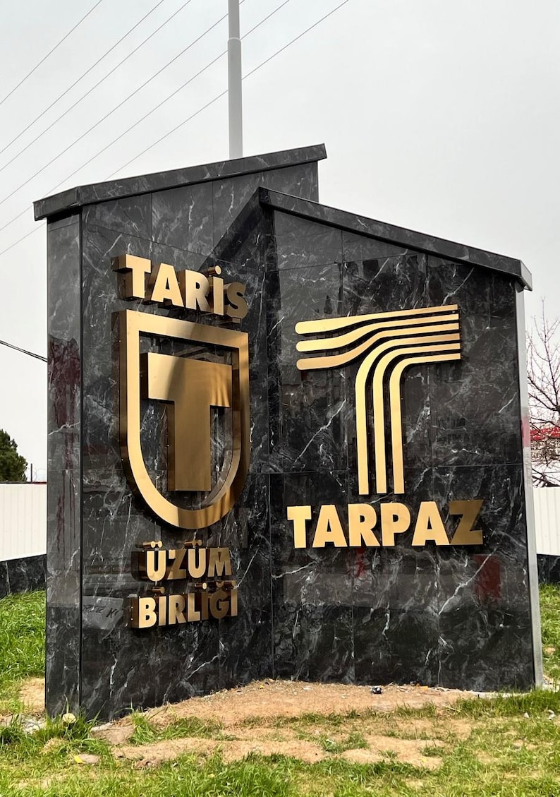 Puede incluir: Un gran monumento angular hecho de m&aacute;rmol oscuro pulido con vetas blancas. Presenta letras y logotipos de metal dorado. El lado izquierdo muestra el texto TARİŞ &Uuml;Z&Uuml;M BİRLİĞİ con un logotipo de escudo, y el lado derecho muestra TARPAZ con un logotipo de T.