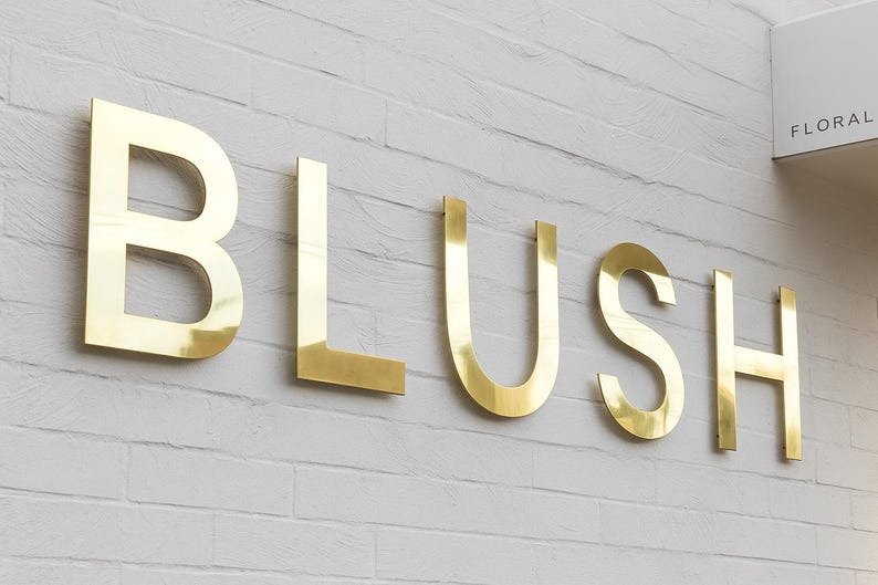 May include: Gold-colored metal letters spelling "BLUSH" are mounted on a white brick wall. The letters are large and shiny, with a modern, minimalist design. A sign that says "FLORAL" is in the upper right corner.