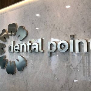 May include: A wall-mounted sign featuring the text "dental point" in metallic silver lettering. To the left of the text is a circular logo composed of six stylized tooth shapes arranged in a ring, all mounted on a white marble surface.
